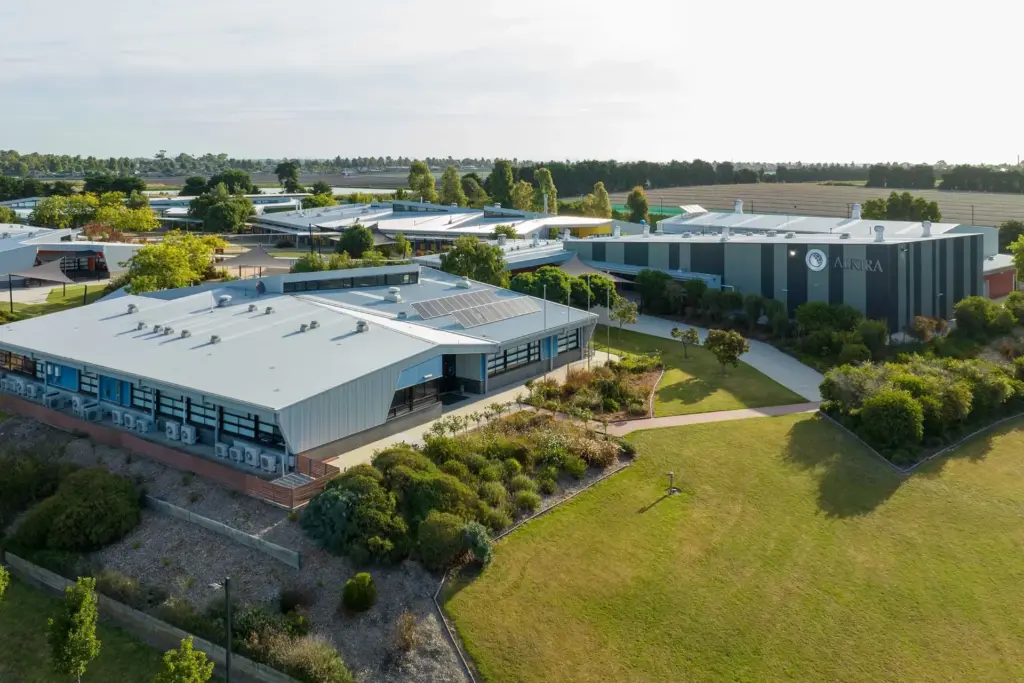 Aerial view of a modern school campus with several low, grey buildings, landscaped gardens, pathways, and open grassy areas, surrounded by trees and open fields.