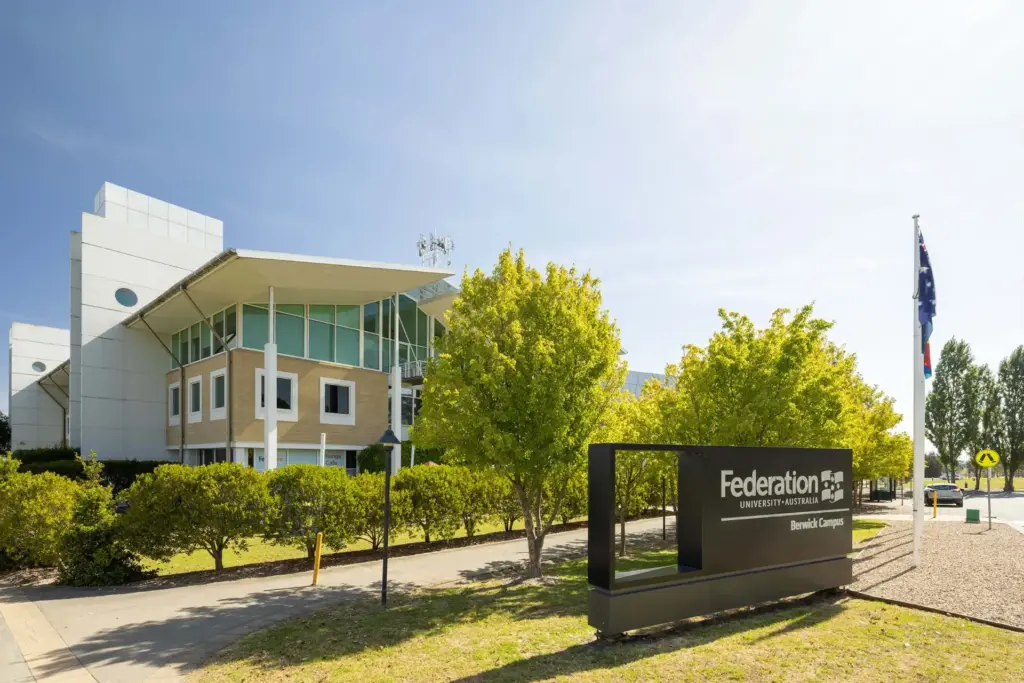 Modern university building with large windows and trees in front. A sign reads "Federation University Australia, Berwick Campus." An Australian flag is visible on the right under a clear blue sky.