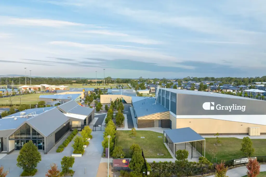 Aerial view of a modern school campus with several buildings, green lawns, and trees. A large building on the right displays the name "Grayling." Houses and open fields are visible in the background under a blue sky.