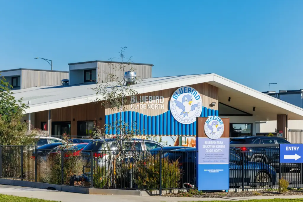 A modern building with a sign reading "Bluebird Early Education Clyde North," featuring the Bluebird logo, parked cars, trees, and a blue arrow sign indicating the entry.