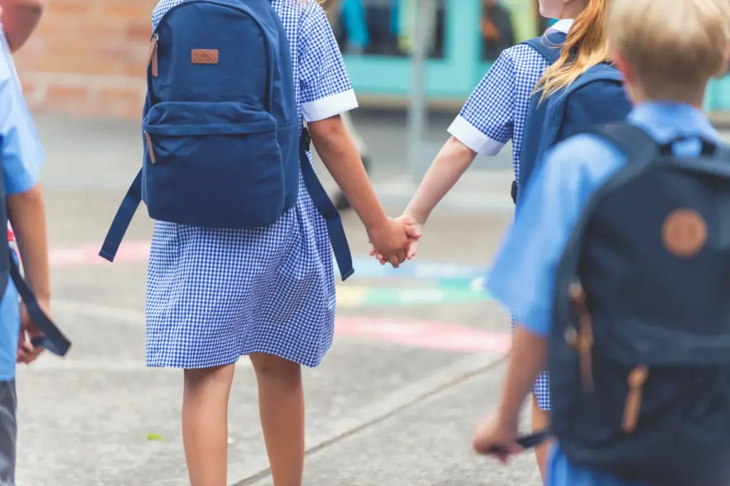 Two young children in blue checkered school uniforms and backpacks hold hands as they walk outside, while other children in similar uniforms walk nearby. The scene appears to be at a school or playground.
