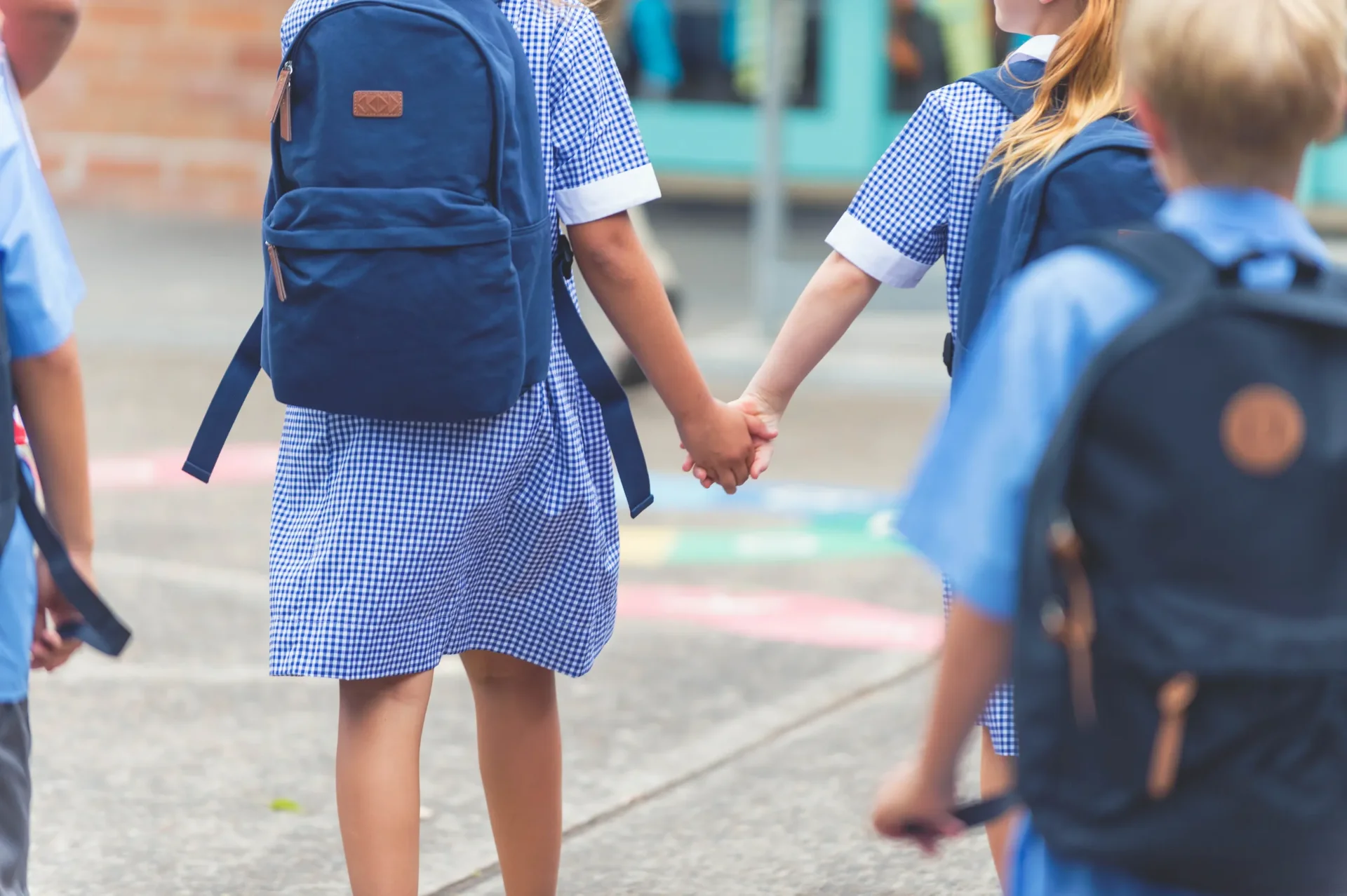Two young children in blue checkered school uniforms and backpacks hold hands as they walk outside, while other children in similar uniforms walk nearby. The scene appears to be at a school or playground.