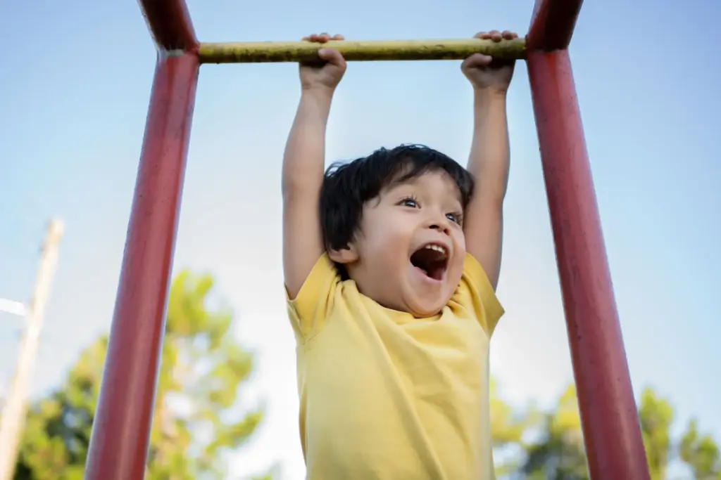 A young child in a yellow shirt hangs from playground monkey bars outdoors, smiling joyfully with an open mouth. The sky is clear and trees are visible in the background.