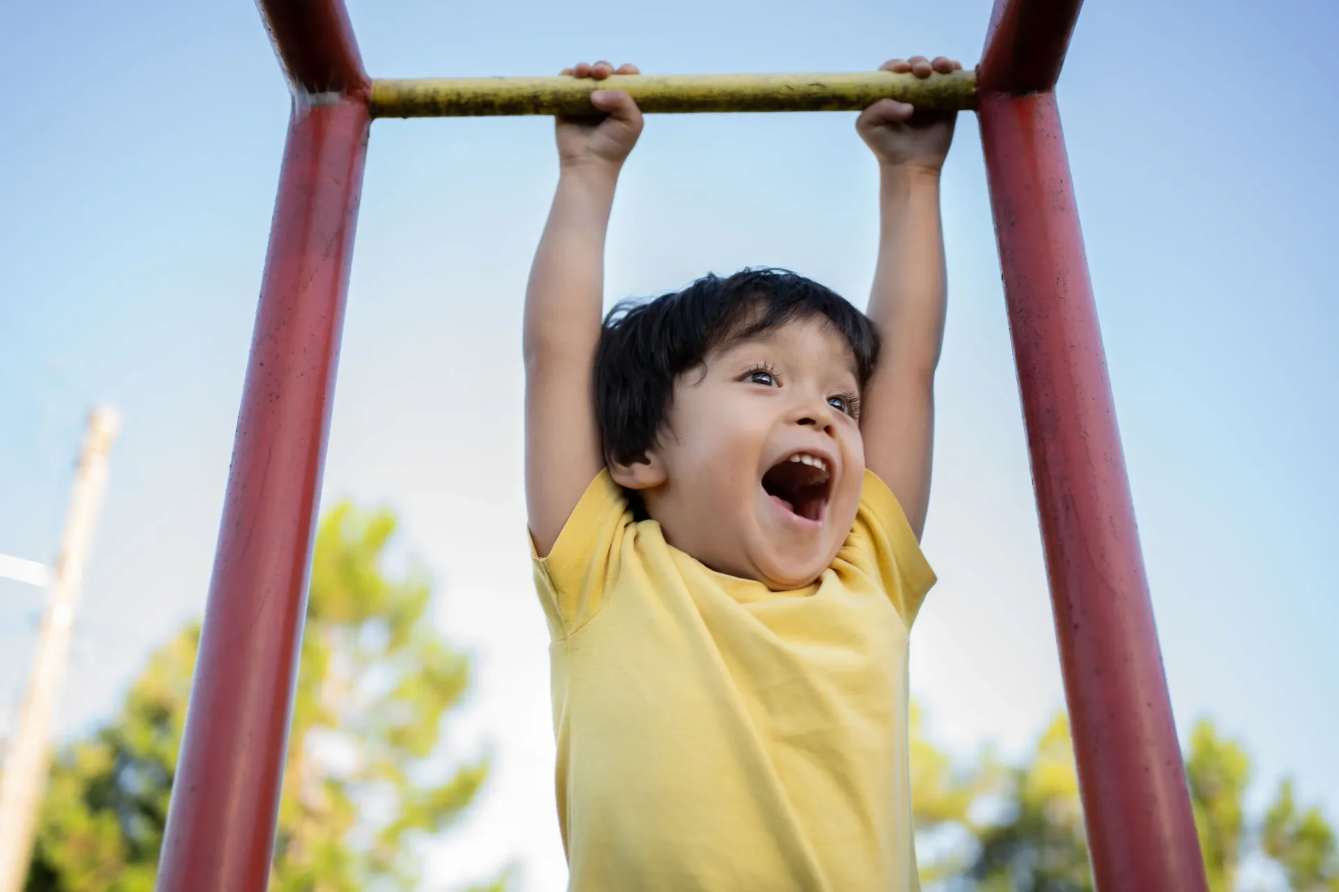 A young child in a yellow shirt hangs from playground monkey bars outdoors, smiling joyfully with an open mouth. The sky is clear and trees are visible in the background.