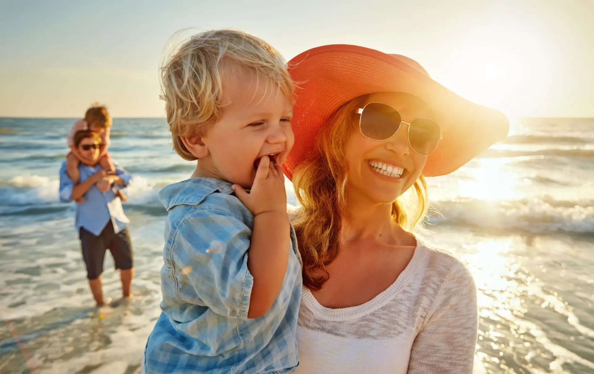 A smiling woman in a sunhat and sunglasses holds a laughing young boy on a sunny beach, while a man carries a child in the background by the water with waves and a bright sunset.