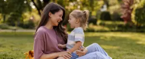 A woman and a young child sit on a yellow blanket in a park, smiling and laughing together. A plush toy is beside them, and trees and greenery are visible in the background.